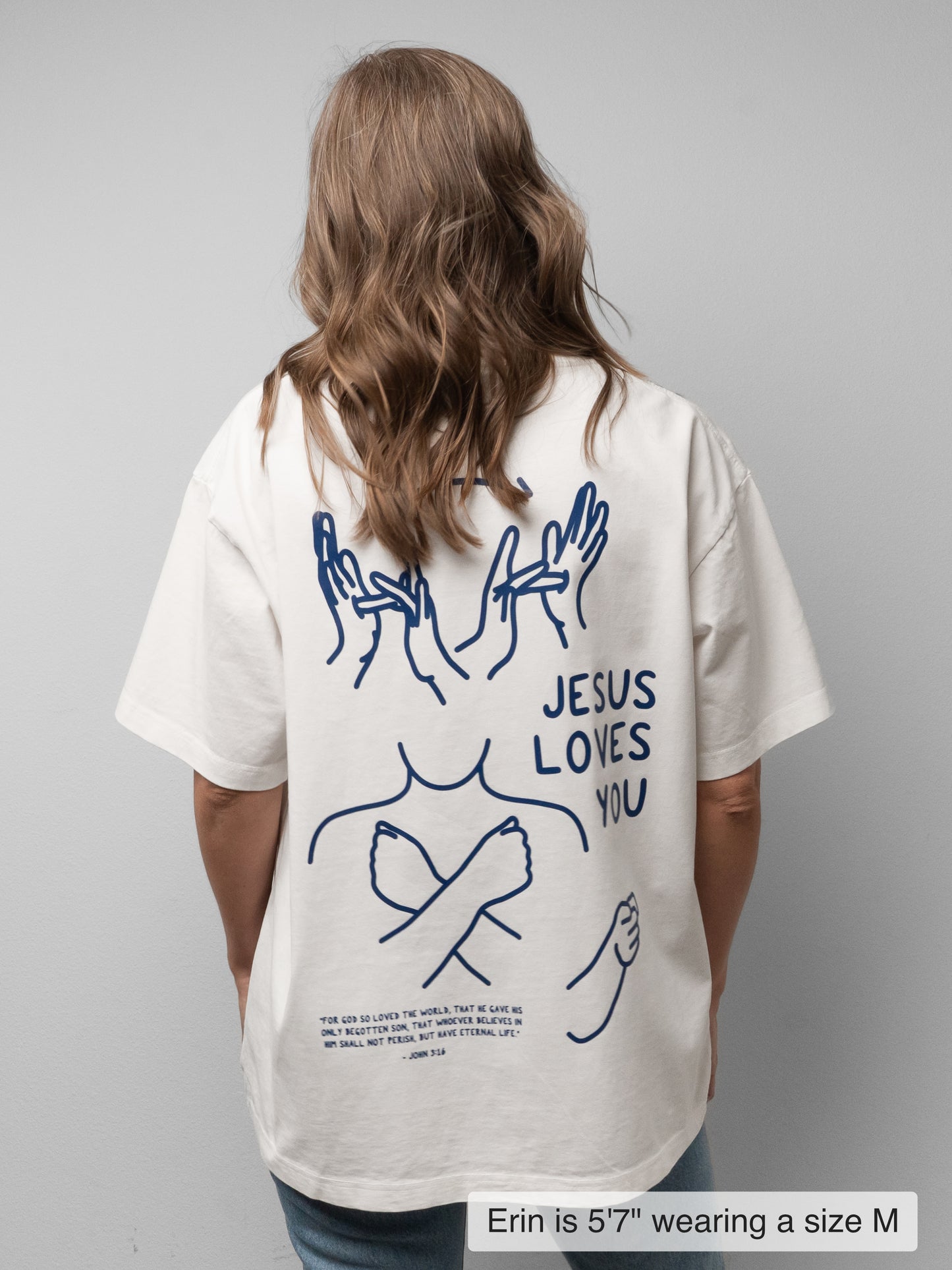 A woman viewed from behind wearing an oversized off-white t-shirt with a blue graphic print on the back. The print features hand illustrations, the text 'JESUS LOVES YOU', and a small Bible verse.
