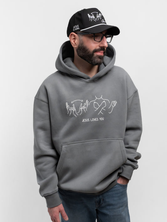 Man wearing a gray hoodie with 'Jesus Loves You' text and a black cap on a white background