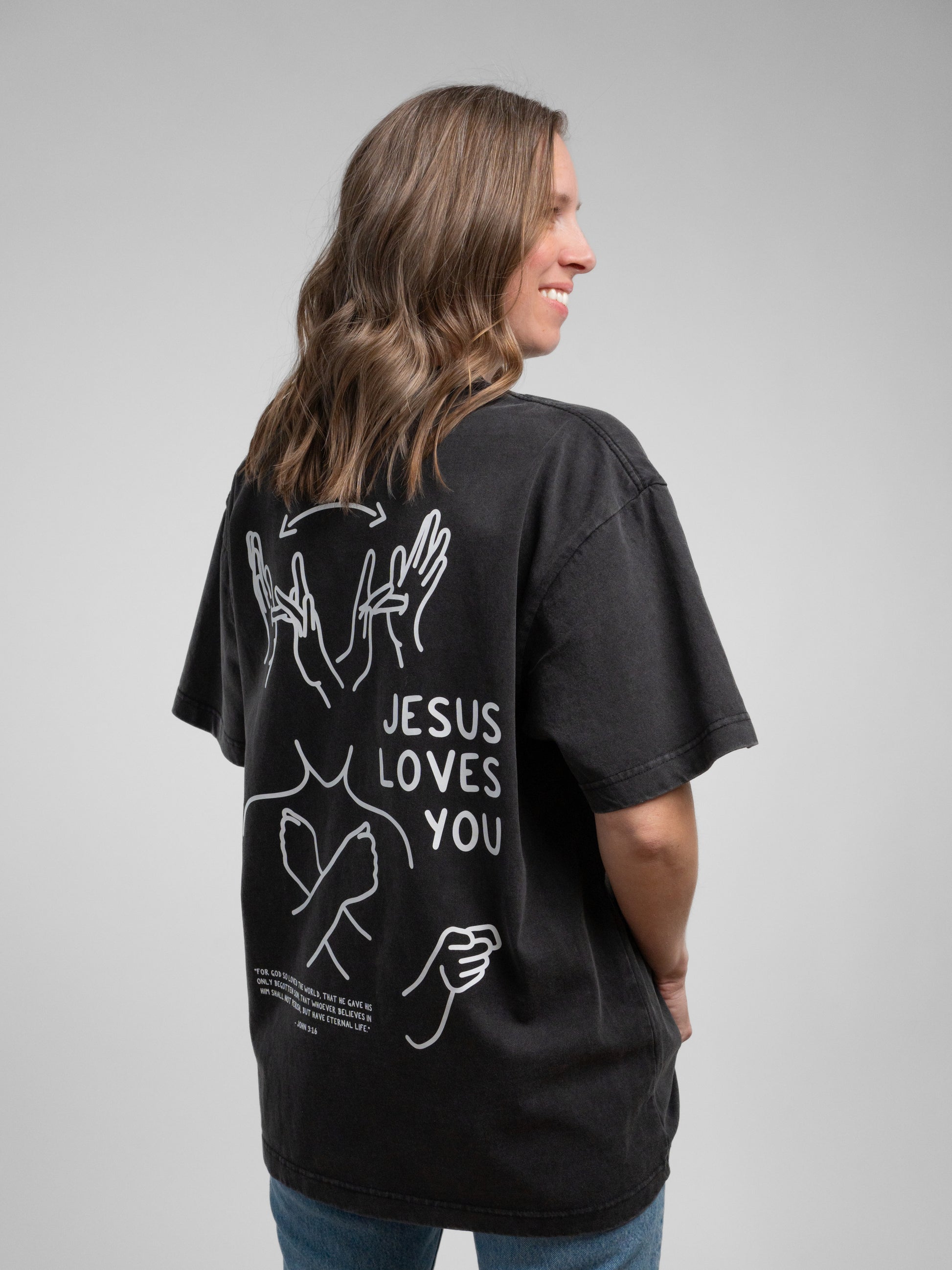 Person wearing a black t-shirt with 'Jesus Loves You' text on the back, standing against a gray background