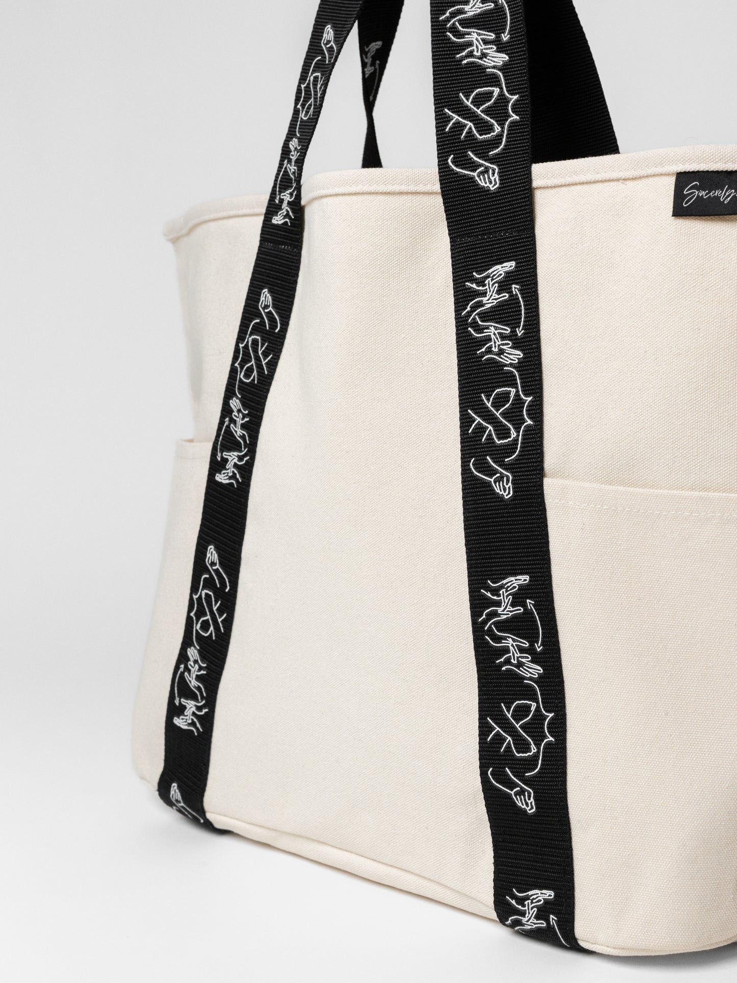 Beige tote bag with black straps featuring white designs on a white background