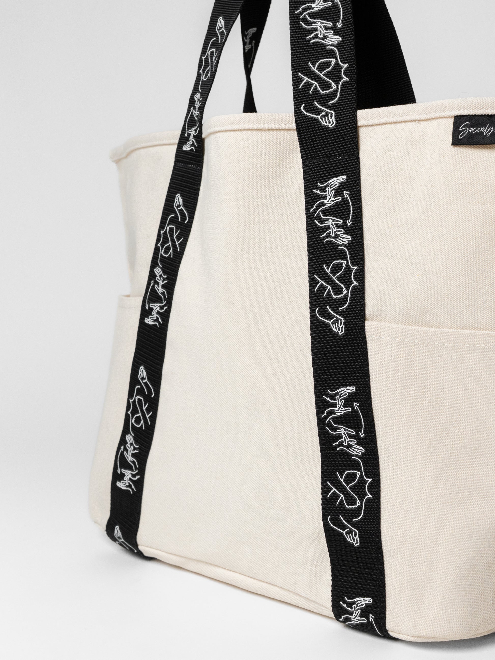 Beige tote bag with black straps featuring white designs on a white background