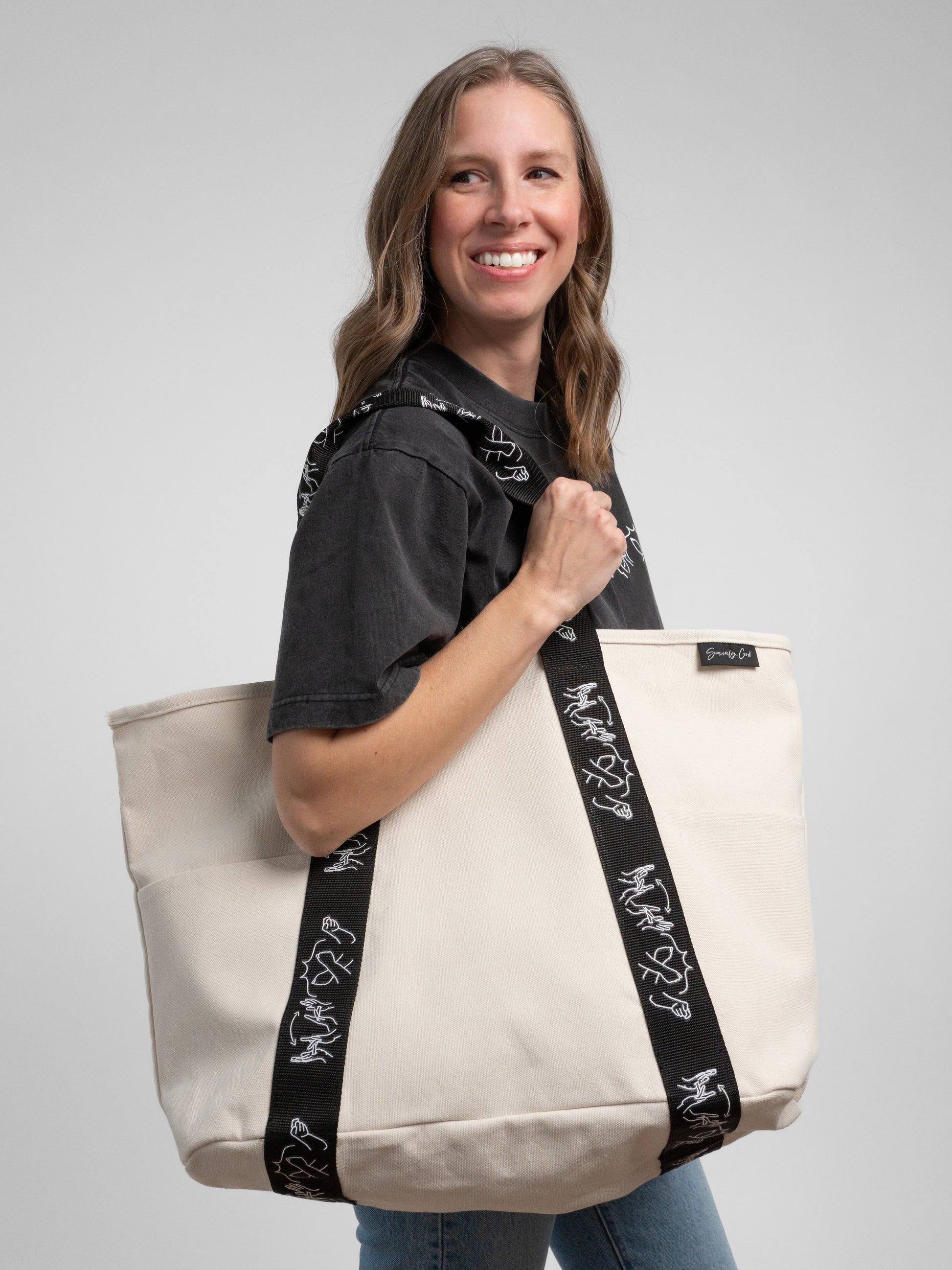 Woman holding a beige tote bag with black straps featuring white designs on a gray background