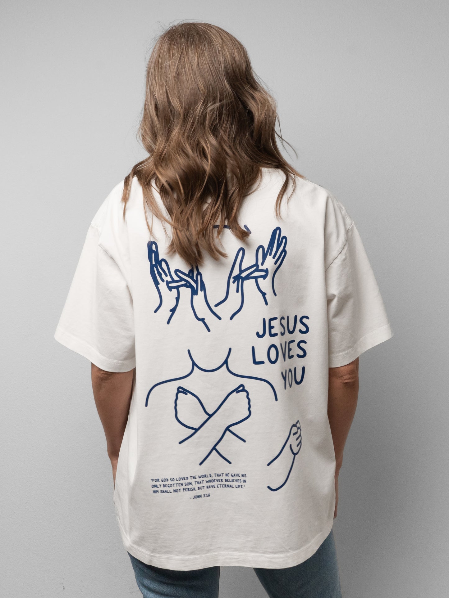 Person wearing a white t-shirt with 'Jesus Loves You' text and graphic design on the back.